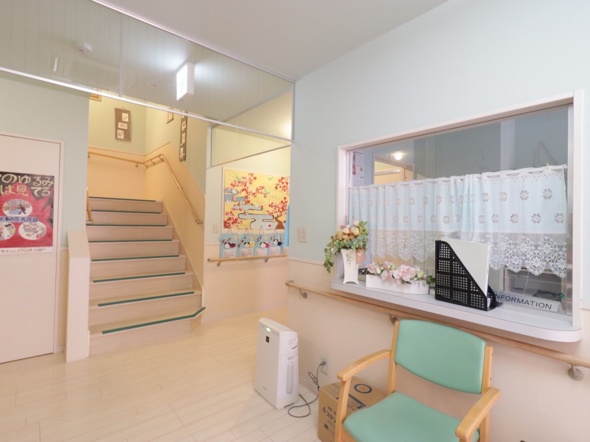 受付のカウンターの上に花が飾られている。ガラス扉があり、カーテンがひいてある。前に椅子が置いてある。階段や通路に手すりが完備されている。