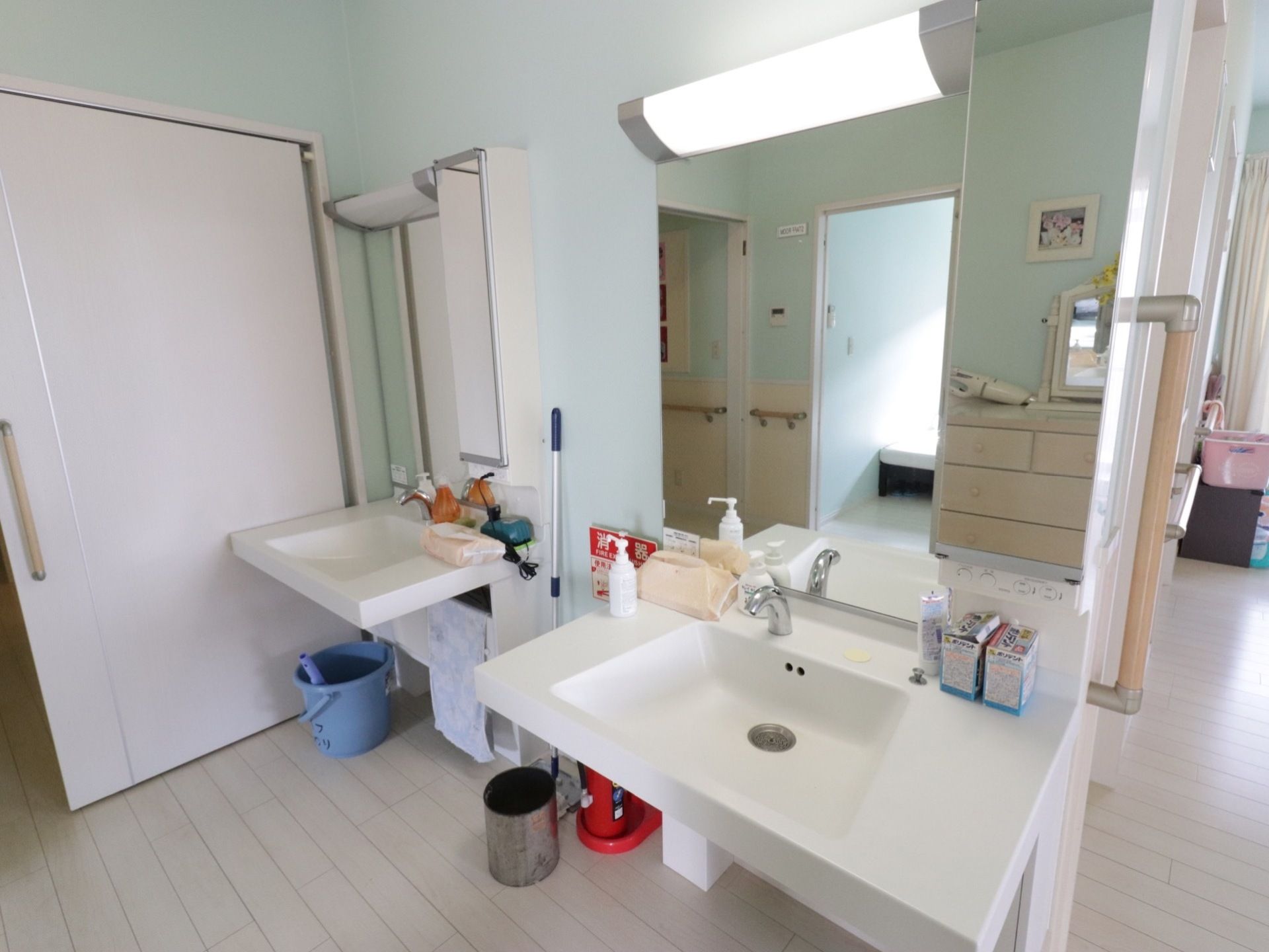 洗面台が２台並んで設置されている。鏡が設けられ、上に照明がついている。ゴミ箱、掃除道具が置かれている。