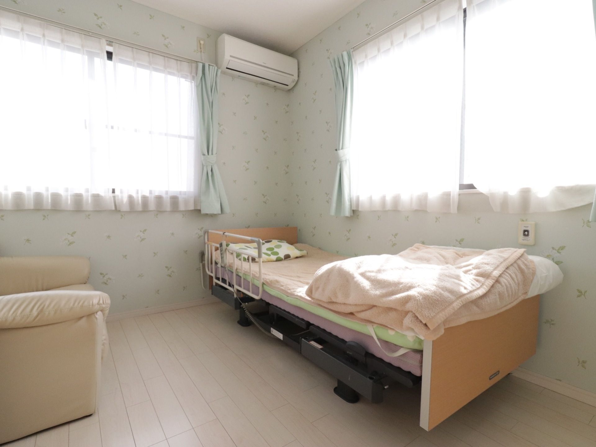 エアコンと窓を2面設置している居室で陽射しが入り明るい。壁紙は爽やかな柄で介護用ベットやソファーを設置している。