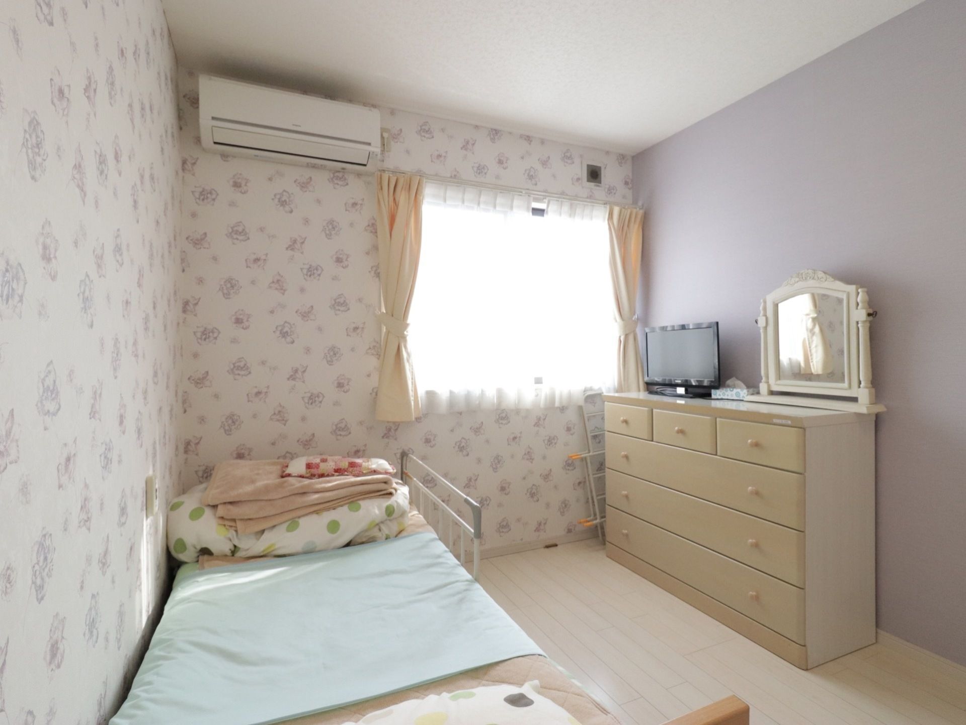 華やかな壁紙を貼った居室である。窓があり介護用ベットやチェストが設置されており、上にはテレビや鏡が置かれている。