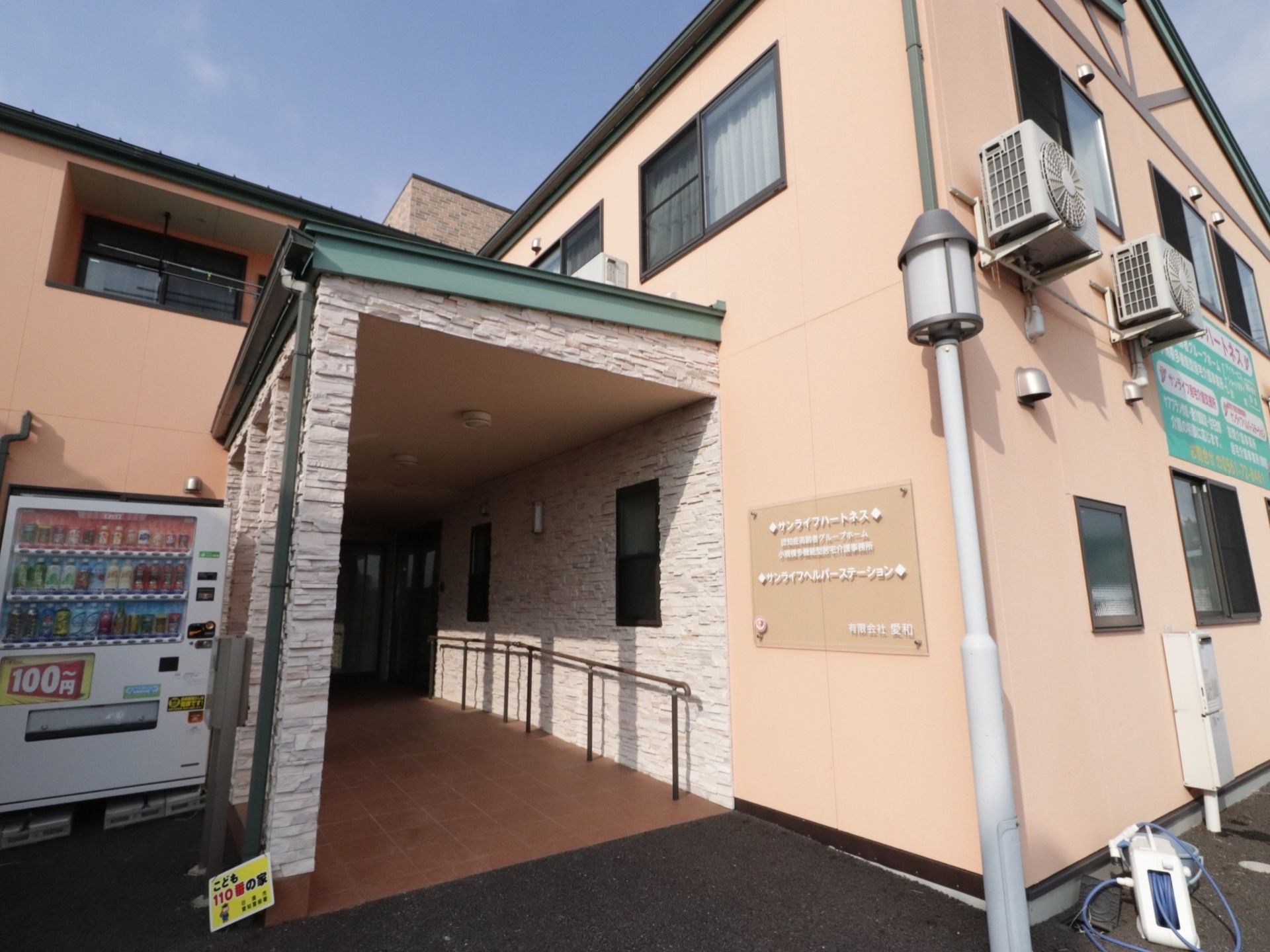 入口付近には外灯を設置し、壁には施設のネームプレートが貼られている。屋根下に手すり付きスロープが設置され、左側には自動販売機がある。