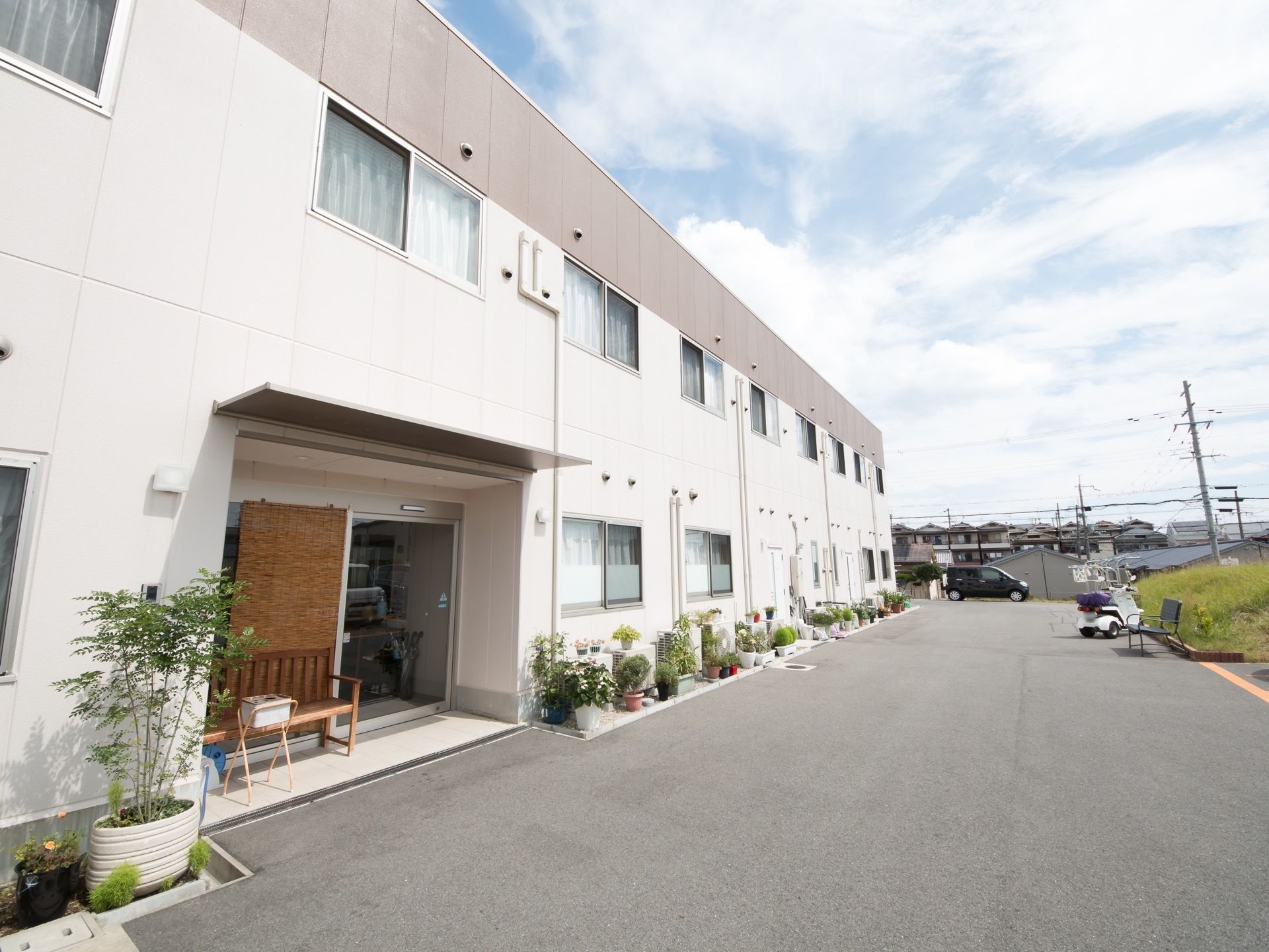 写真の奥には住宅が軒を連ねているのが見える。施設のそばには、黒い乗用車とスクーターが停められている。