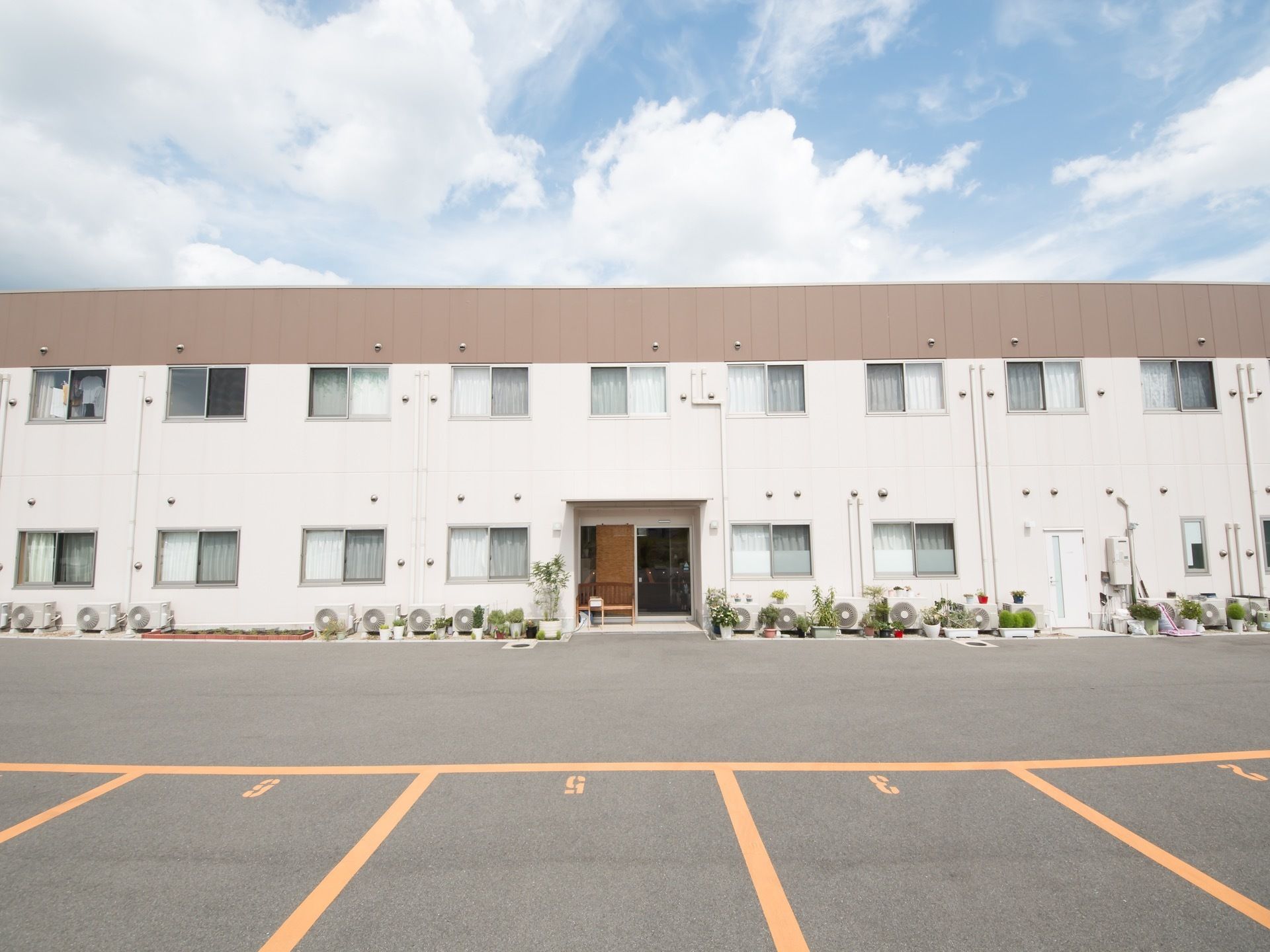 白い壁の２階建ての建物である。建物の前には室外機とさまざまな植物の鉢植えが置かれている。施設前は駐車場が広がっている。
