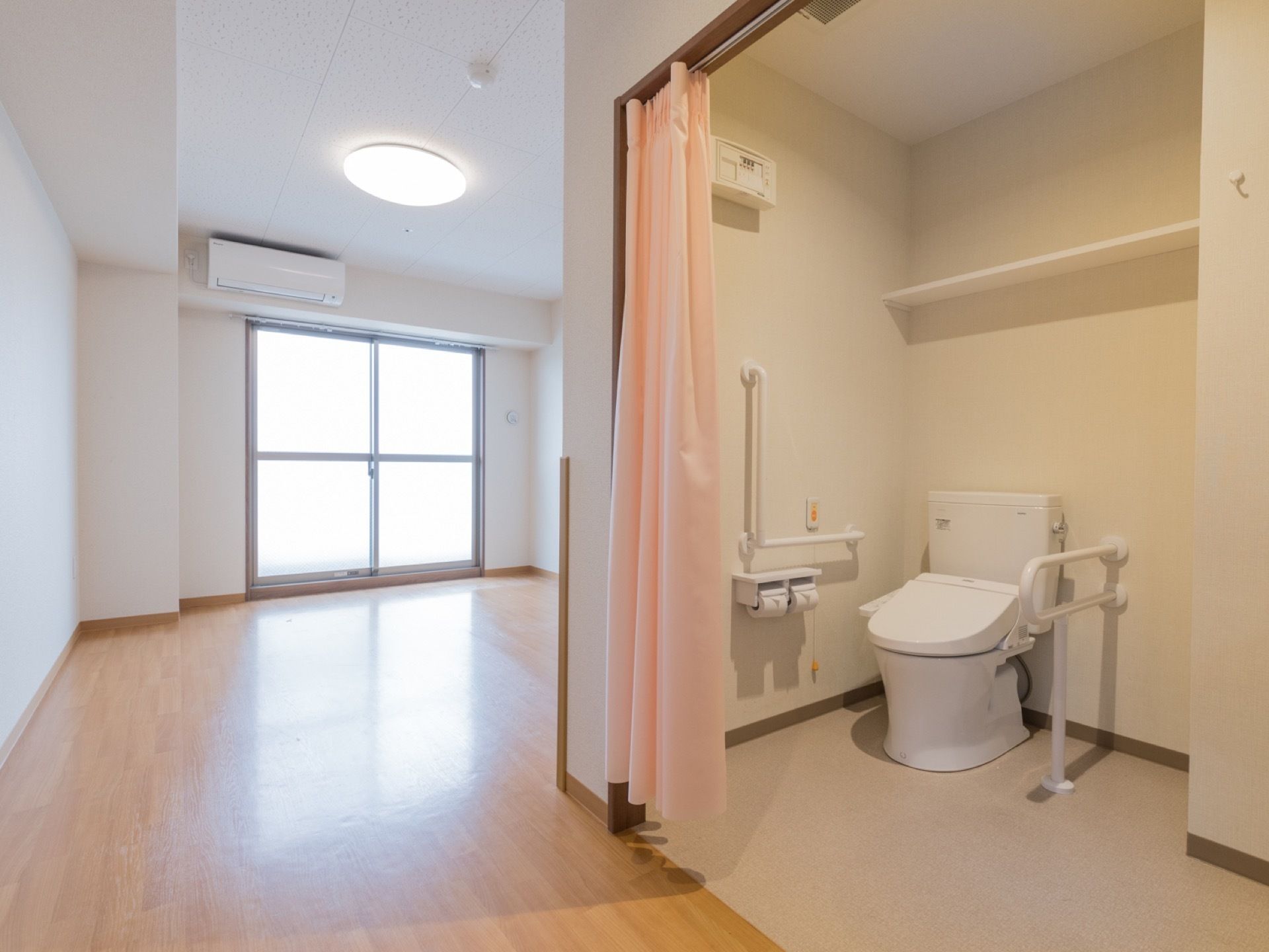 大きな窓から自然光が差し込む明るい居室である。右端にはトイレがあり、ピンク色のカーテンで仕切りができるようになっている。