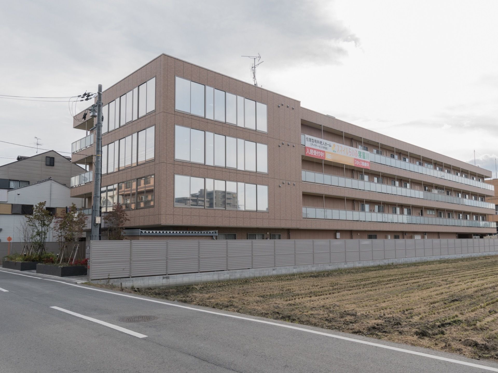斜め方向から、建物の全体像を写したものである。茶色でまとめられた落ち着いた外観となっている。右横は田んぼのような空地である。