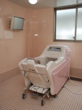 薄いピンク色を基調とした介護浴室である。キャスターのついた椅子が浴槽に備え付けられており、壁にはテレビパネルが設置されている。
