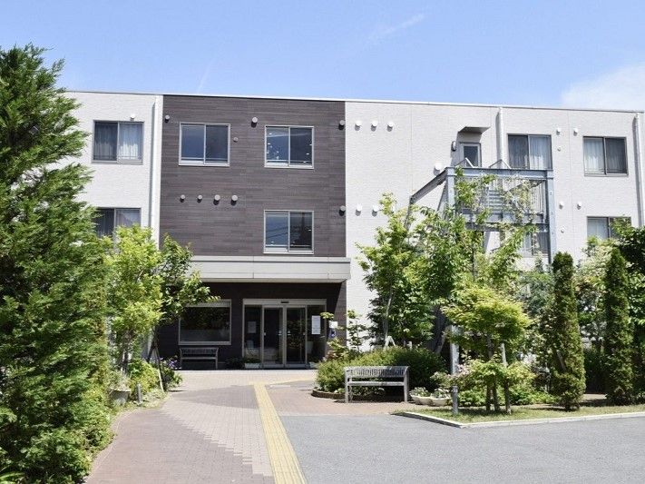 施設は３階建ての落ち着いた印象の建物で、手前には駐車場がある。道路に向けて、施設名を表記した表札と看板がある。