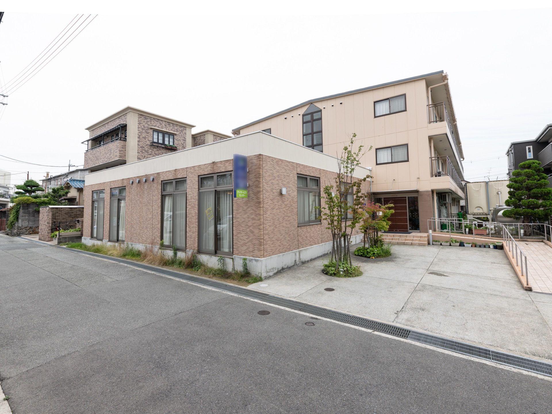 施設は広い道路に沿って建っている。手前にデイサービスセンターがあり、奥に住宅がある。住宅は３階建てで、玄関までスロープがついている。