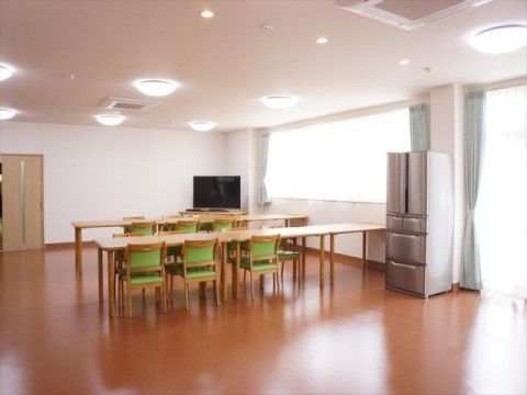 白の内装にフローリング、正面奥にテレビ、その前につなげたテーブルが2列セットされた共用スペース