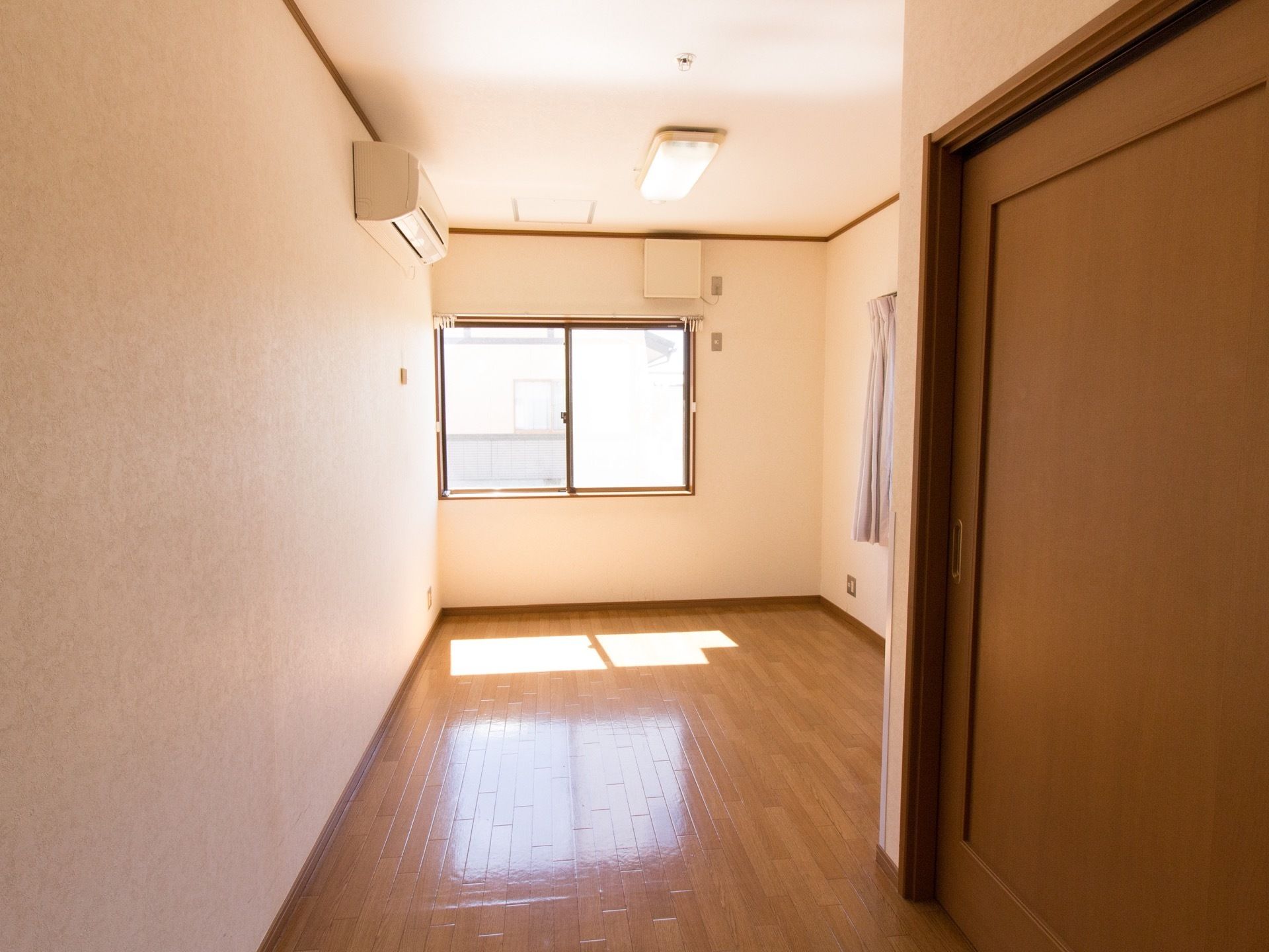 居室の奥には腰高窓があり、外光が降り注いでいる。床が白く輝いている。左側の壁には、エアコンが設置されている。