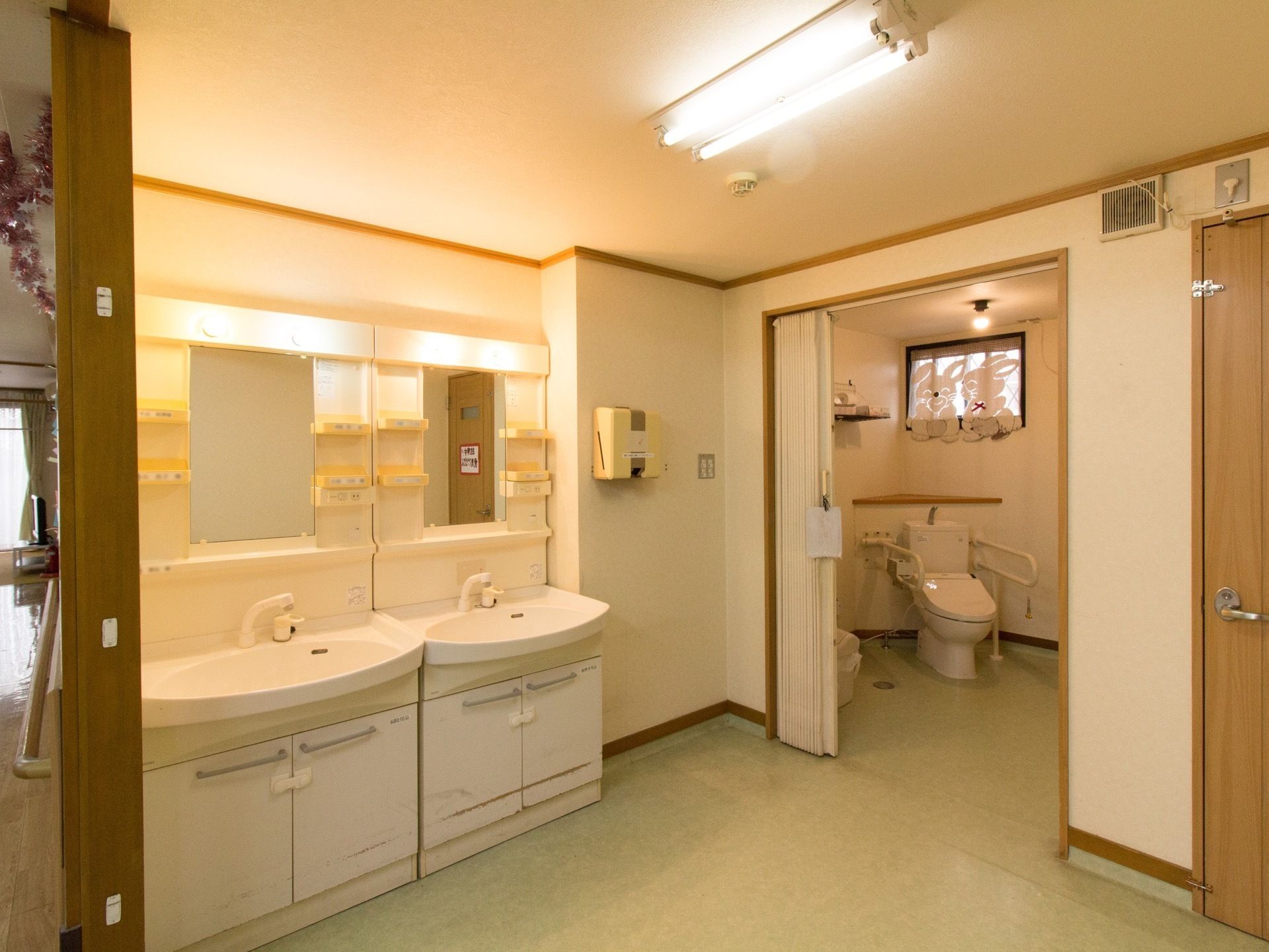カーテンに仕切られてトイレが設置されており、両サイドに手すりがある。白を基調とした洗面台が２つ並んで置かれている。
