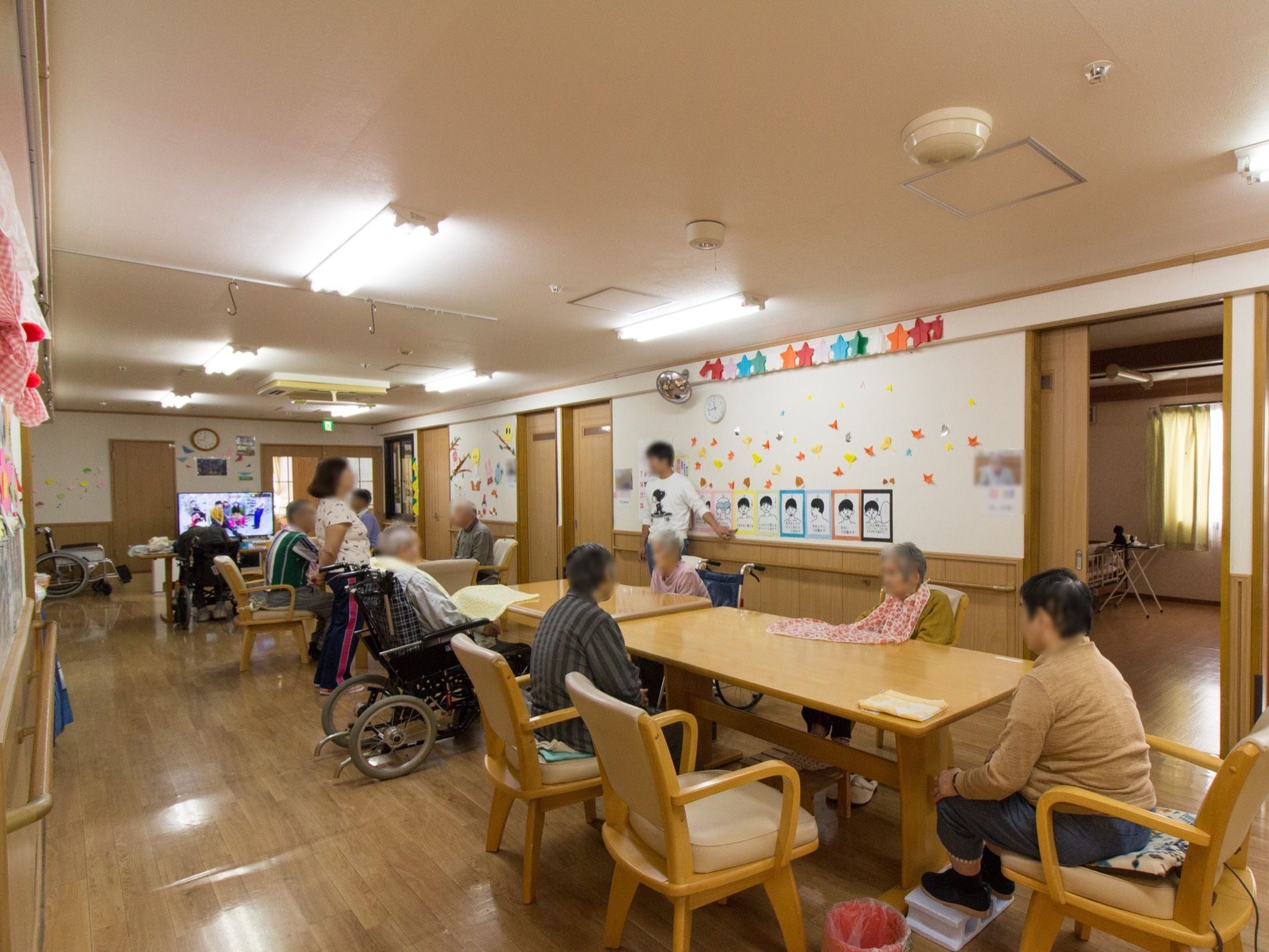 手作りの装飾が施されたリビングで、壁際にスタッフが立っている。大きなダイニングテーブルを囲んで入居者様が座っている。