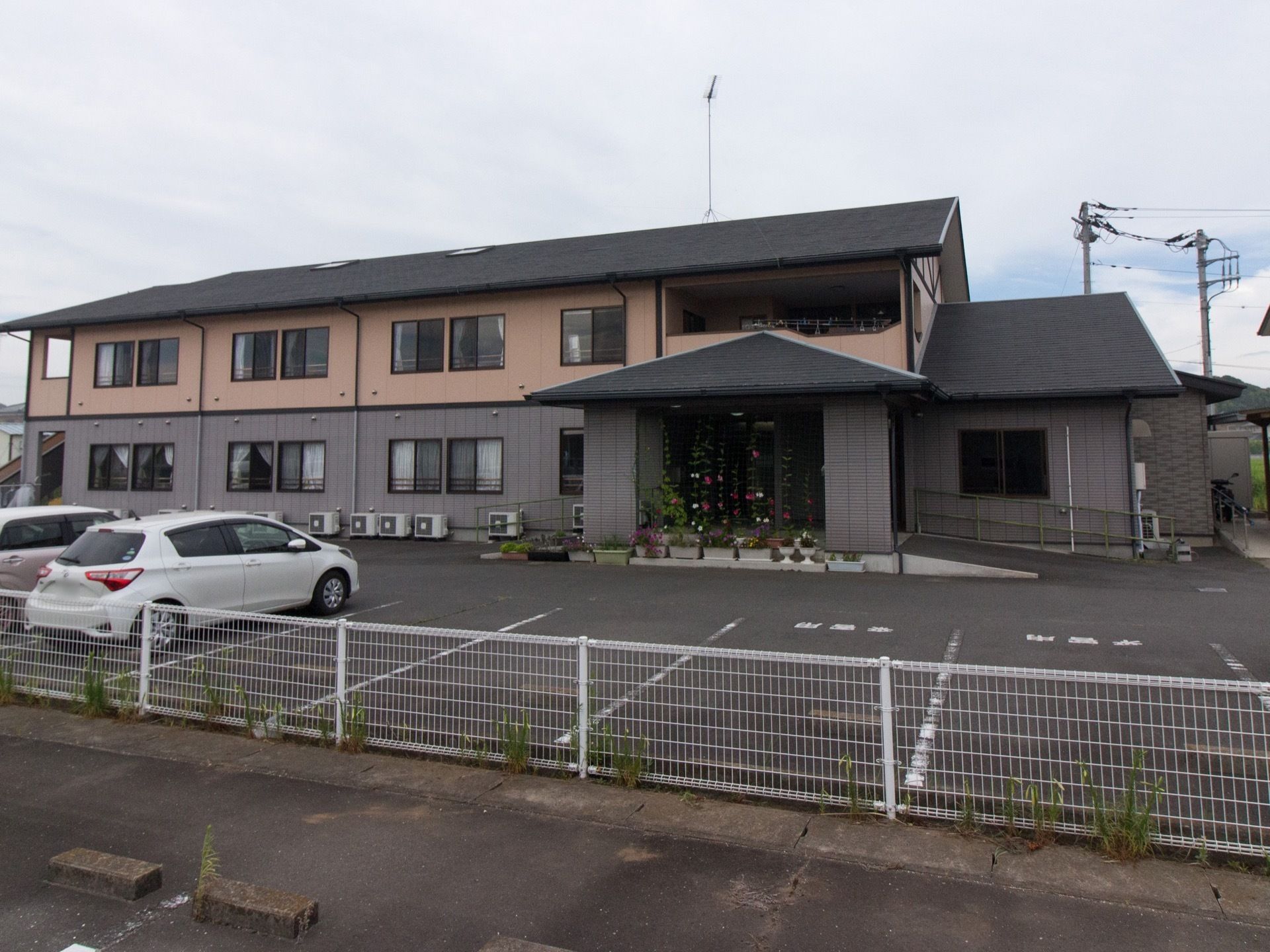 車寄せのある玄関が印象的な、落ち着いた色合いの２階建ての施設である。駐車スペースも十分に確保されている。
