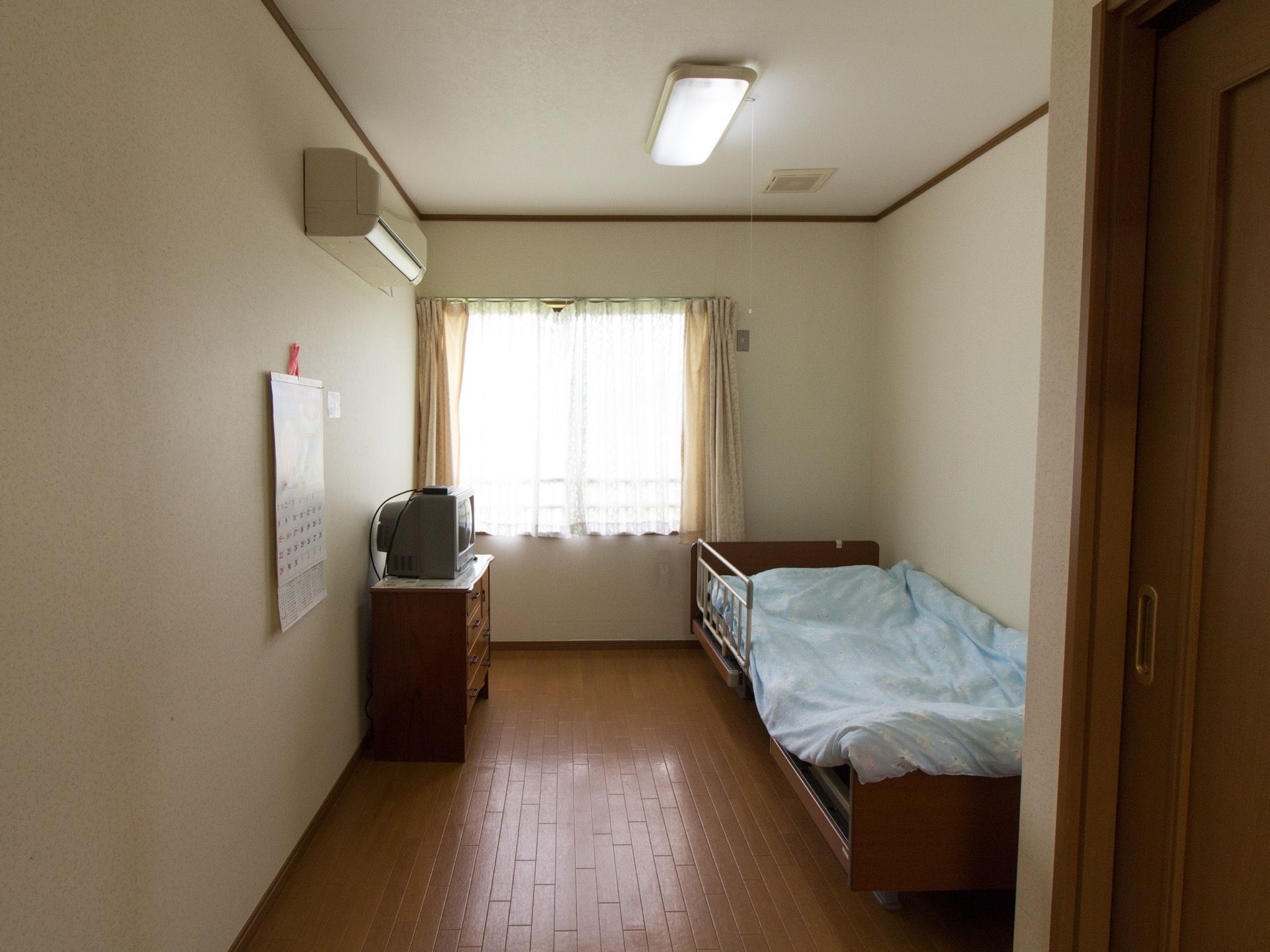 フローリングタイプの居室である。ベッドやテレビ、テレビ台兼収納家具が置かれている。エアコンも設置されている。