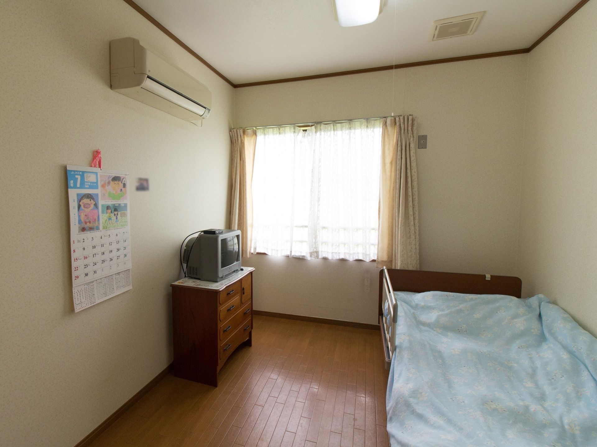 居室は大きな窓からの採光で十分に明るい。ベッドやエアコンが置かれている。テレビ台を兼ねた家具も置かれている。