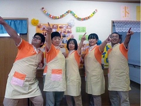 白い壁に掲示物がある場所で、指を伸ばすポーズで立つ5人のスタッフ