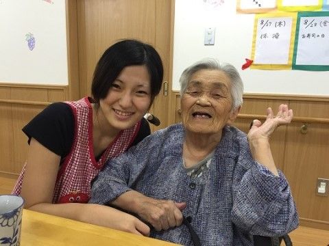 左手を上げて笑顔の白髪の高齢女性とその左に寄り添う女性スタッフ