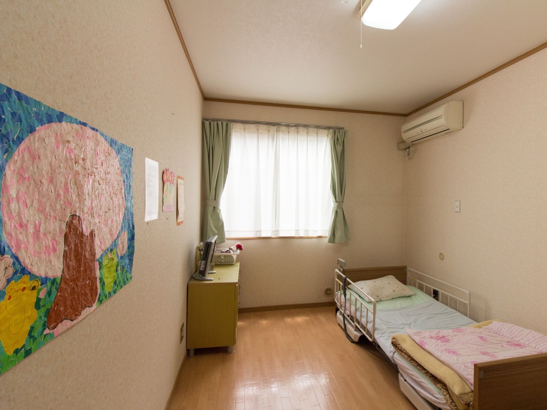 桜の木をモチーフにしたカラフルな貼り絵が、左側の壁に貼ってある。窓際には、モスグリーンの収納ボックスが置いてある。