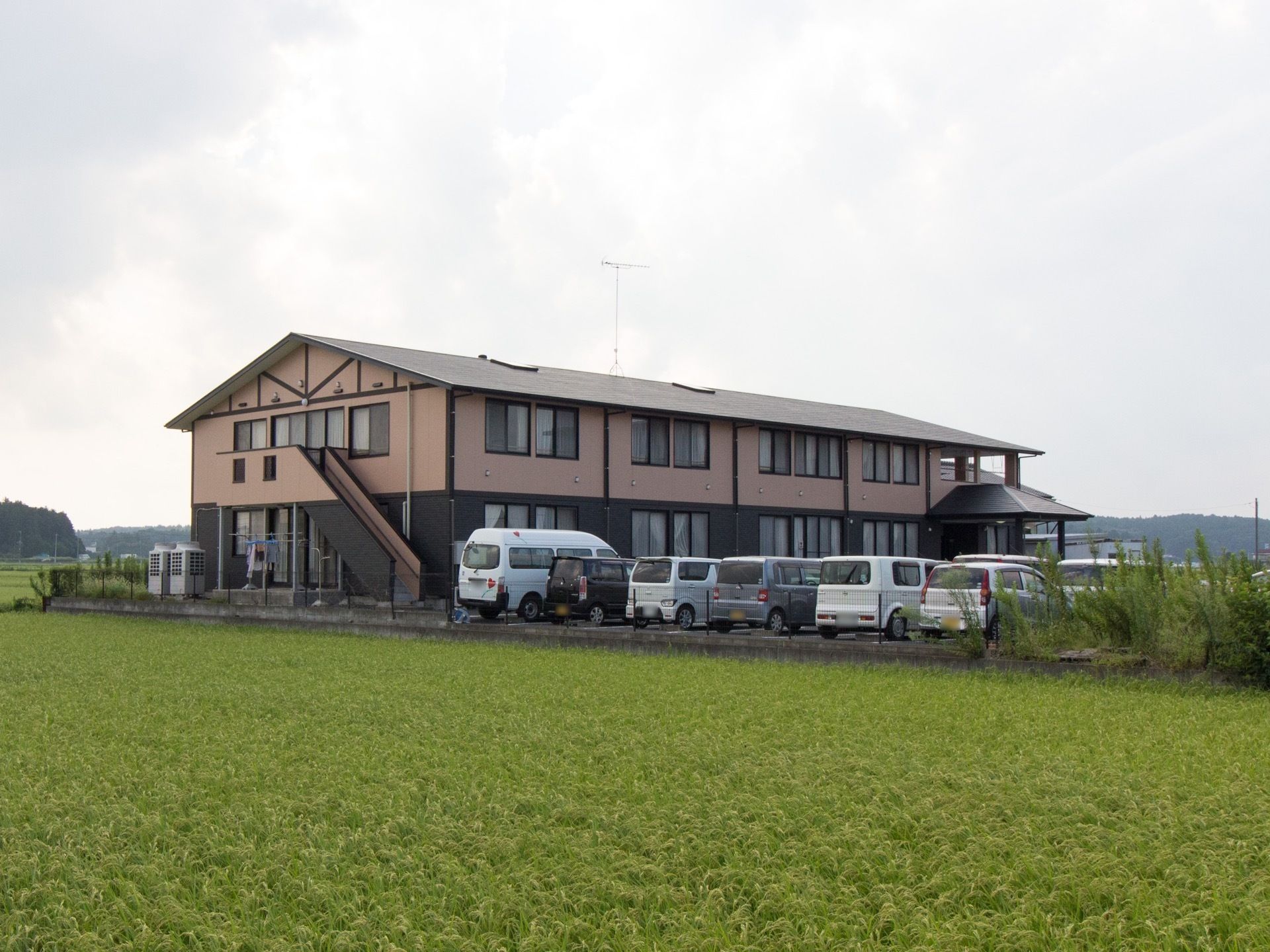 黒と茶色を基調としたモダンな外観の建物。田園風景の中に建っており、建物の前には駐車場があるのが見える。