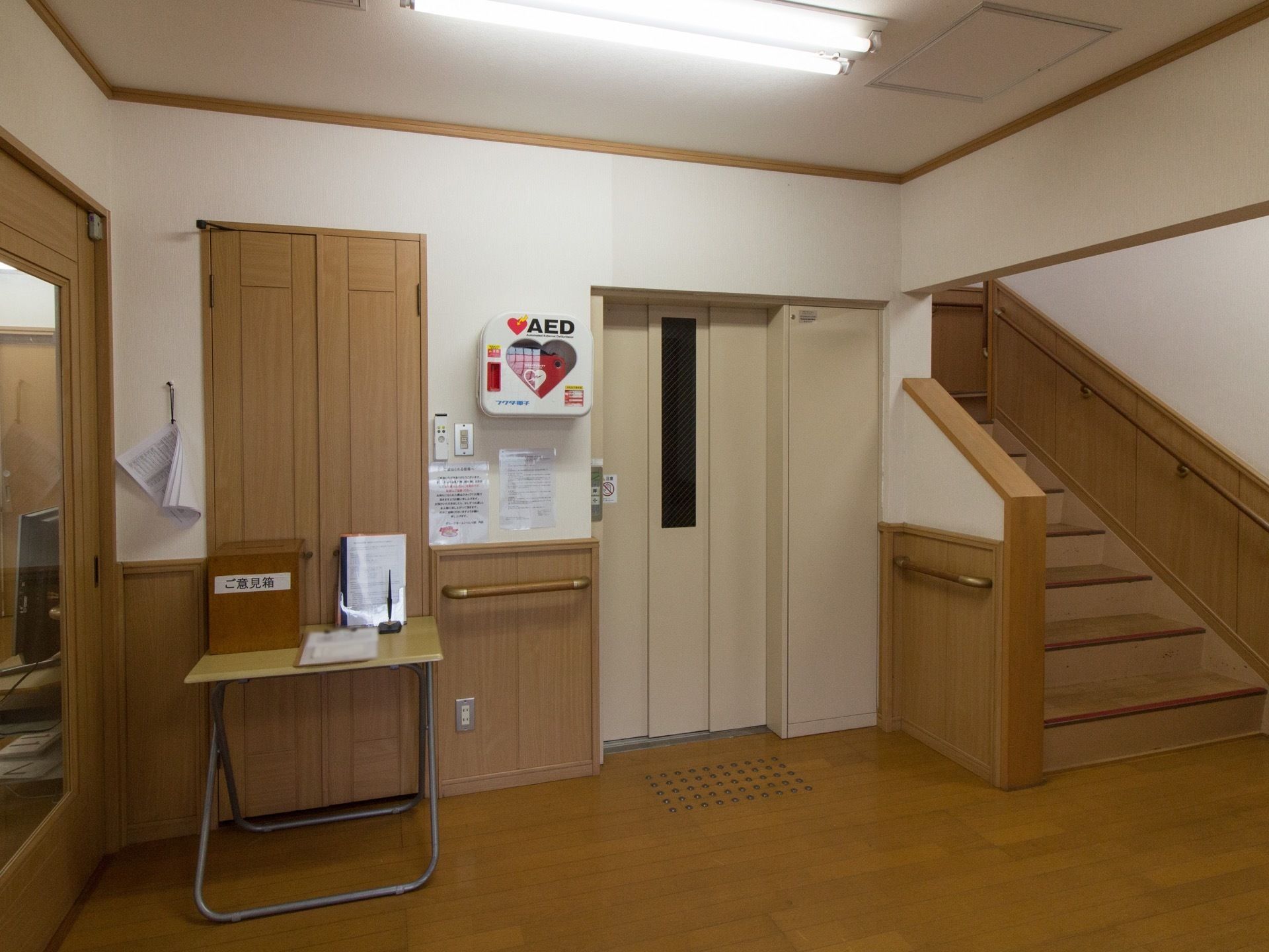 白と茶色の壁に茶色の床の館内。階段の横にクリーム色のエレベーターの扉が見える。壁には手すりが設置されている。