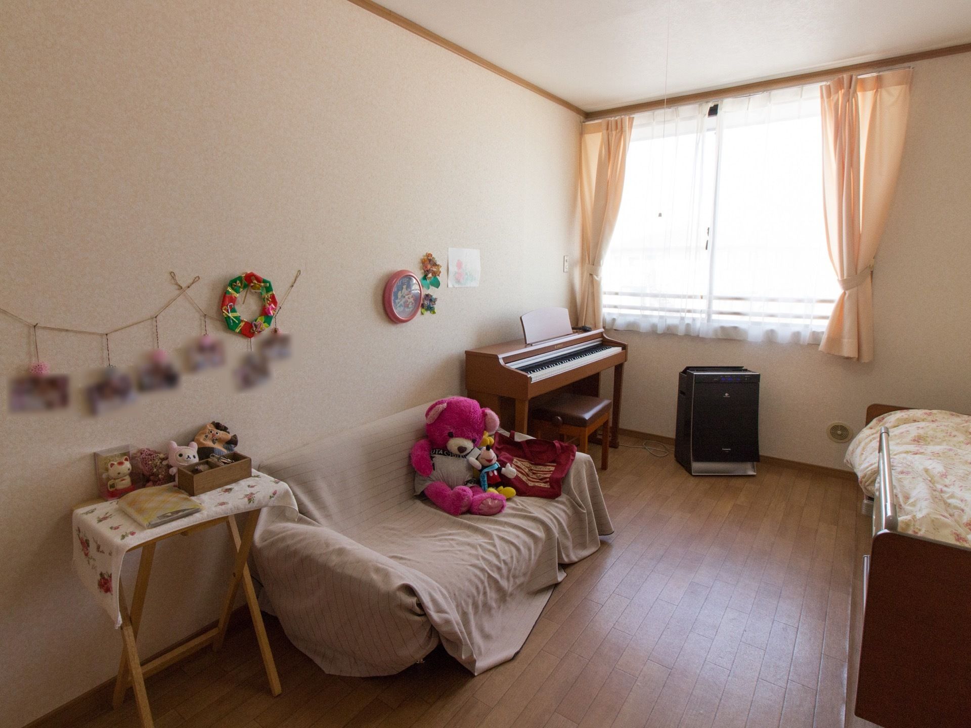 白い壁に木目の床の居室。窓があり日差しが差し込んでいる。ベッドやピアノ、ソファなどが置かれ、ぬいぐるみなどが飾られている。