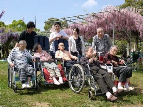 藤棚の前で車いすを利用している5人の高齢者とその後ろに立つ4人の人物
