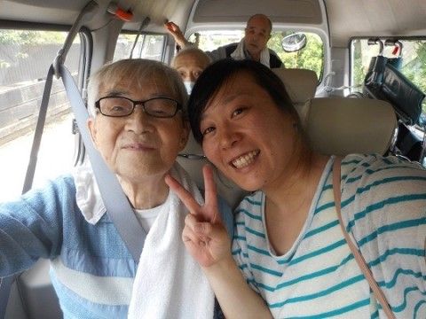 車の中でシートベルトをしている高齢者とその右でピースサインをしているスタッフ