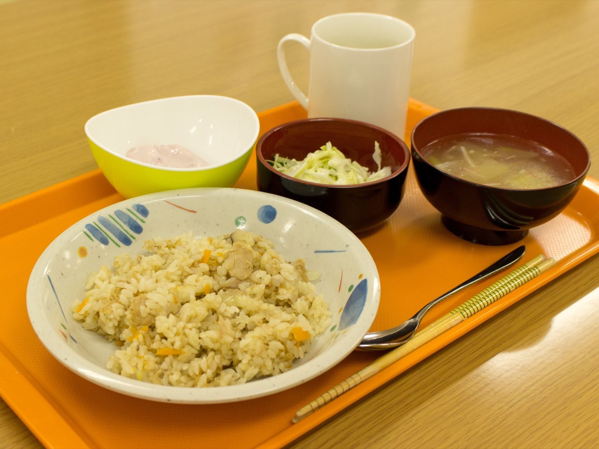 オレンジ色の四角いトレーにそれぞれの皿に盛り付けられた料理が並べられている。箸とスプーンと白いマグカップが置いてある。