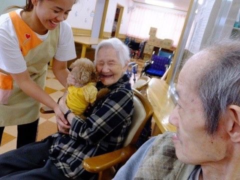 椅子に座って犬を抱く高齢女性と、それを左から介助するスタッフ