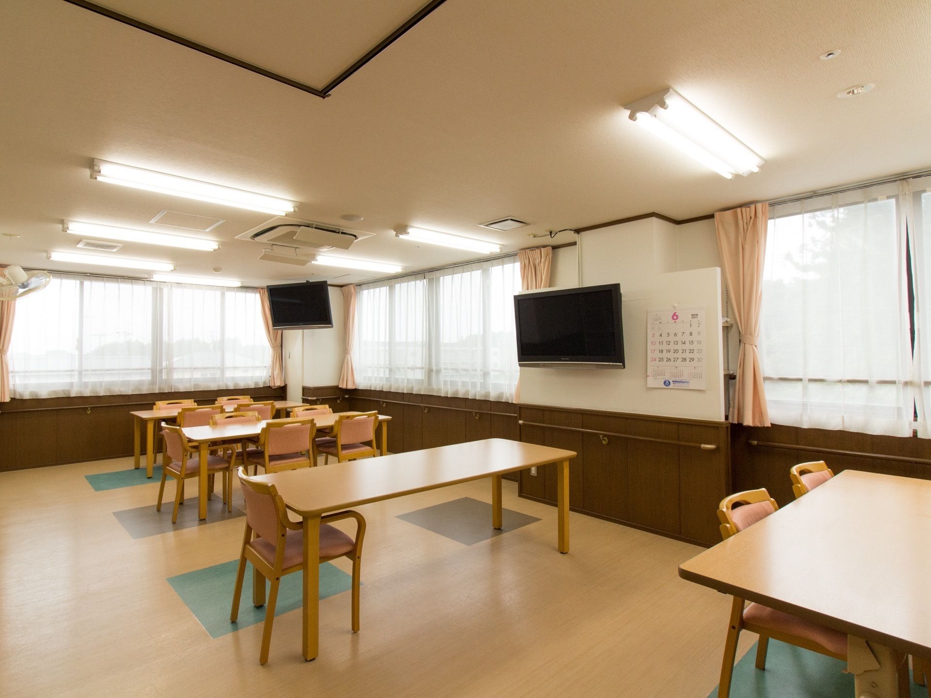 明るく広い共有スペースである。窓が多くとても明るい。手すりがぐるりと設けられており、壁掛けのテレビが2台設置されている。