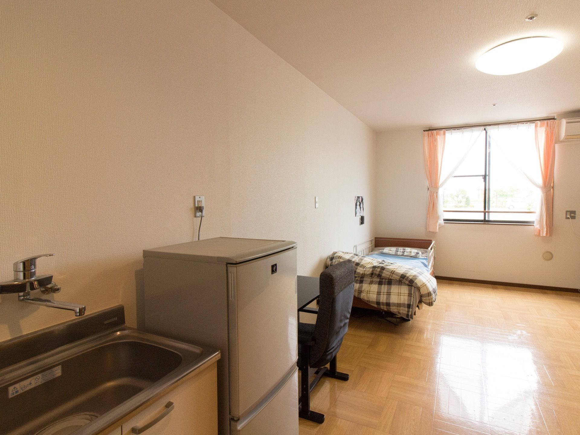 居室は白い色でまとめられている。突き当りに腰高窓がある。天井には丸いシーリングライトがあり、壁際に奥からベッド、机、冷蔵庫、シンクがある。