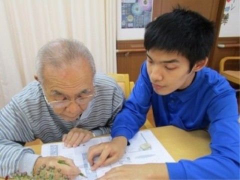 テーブルに広げたものに何か書きこんでいる高齢者と、その右で介助をしているスタッフ