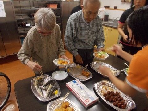 茶色のテーブルの上に置かれた銀色の大皿から手に持った小皿に料理を取り分けている二人の高齢者