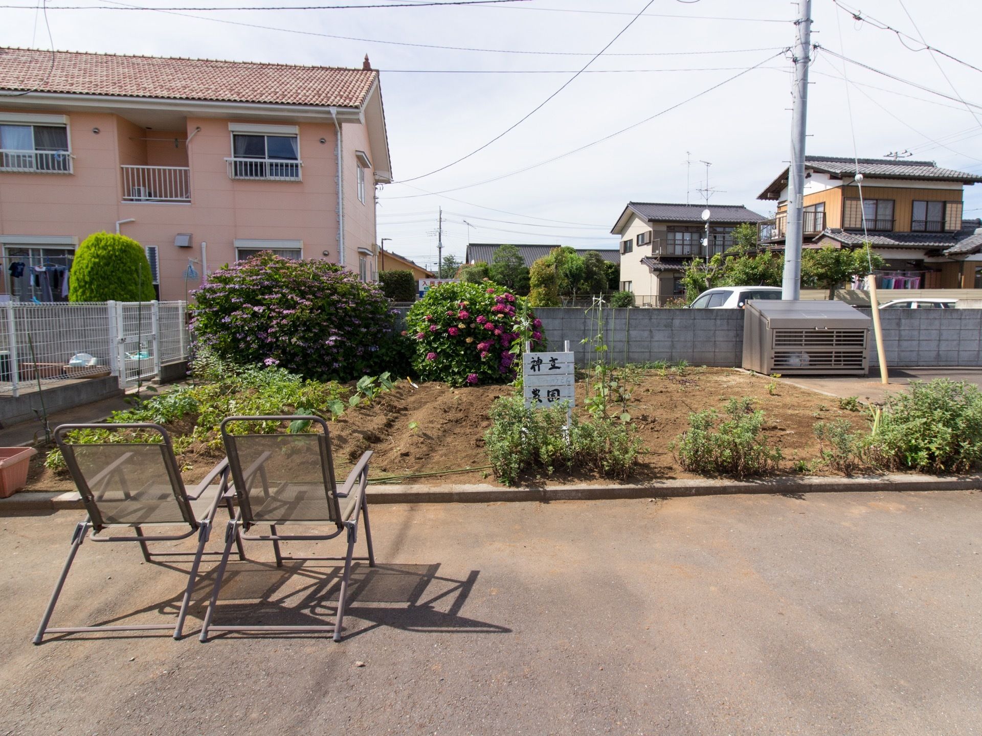 敷地内に日当たりの良い畑があり、白い看板が立っている。畑には農作物や植物が植えられている。手前に二つのデッキチェアが並んでいる。