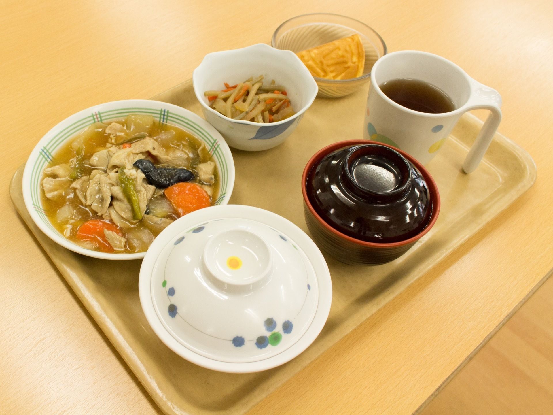 明るい木目調のプレートに食事の一例が並べられている。主食や汁物には蓋をのせ、食べ応えのあるメニューでデザートが添えられている。