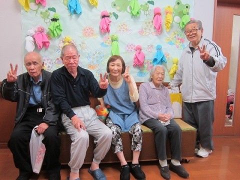 テルテル坊主の掲示物のある壁の前で椅子に座る4人とその脇に立つ1人の人物