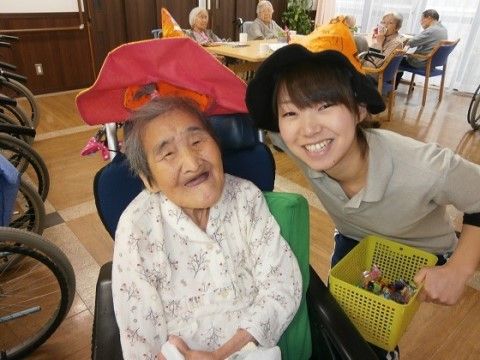車椅子を利用している白いブラウスの高齢女性の右側で仮装の帽子をかぶせようとしている女性スタッフ