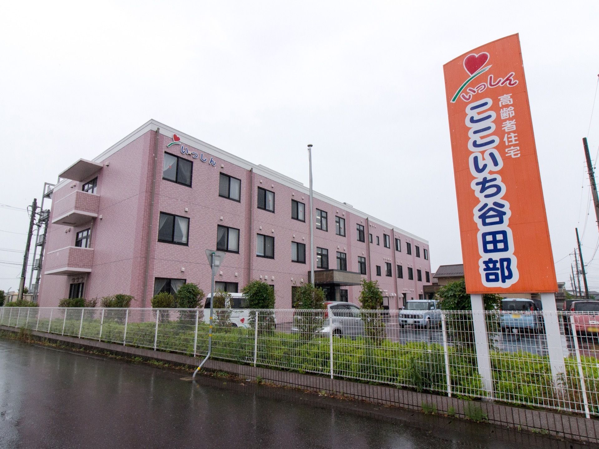 建物はピンクに塗られた３階建てになっている。施設前は駐車場があり、建物の３階部分に大きく看板が取り付けられている。