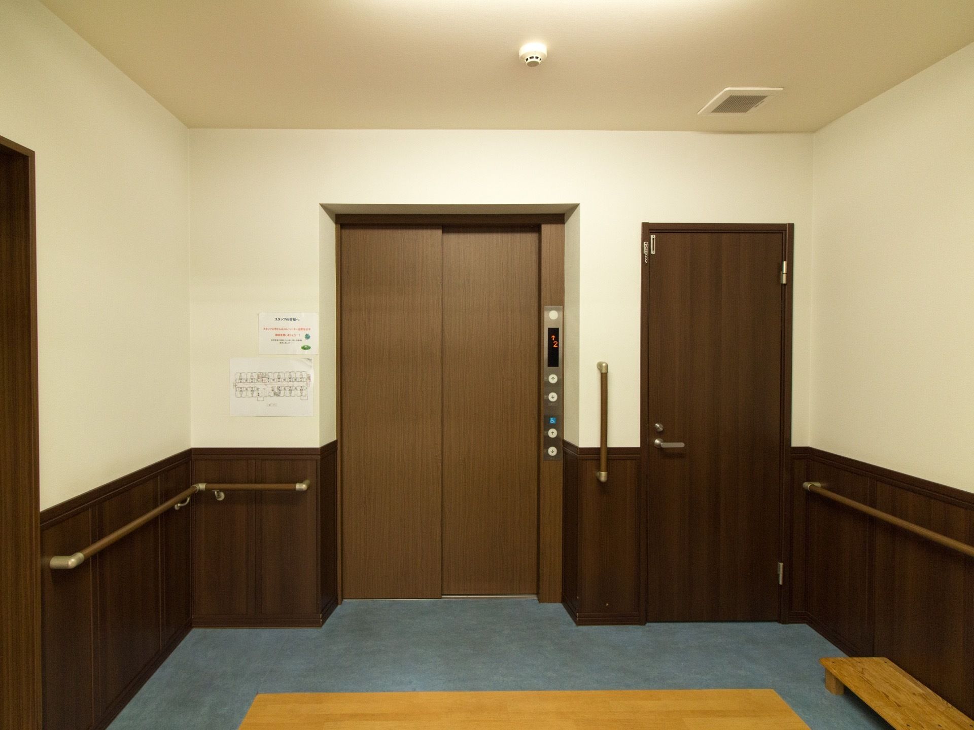 エレベーター横の壁には施設内の案内図が貼られている。壁には手すりが取り付けられている。スタッフ用倉庫の扉がある。