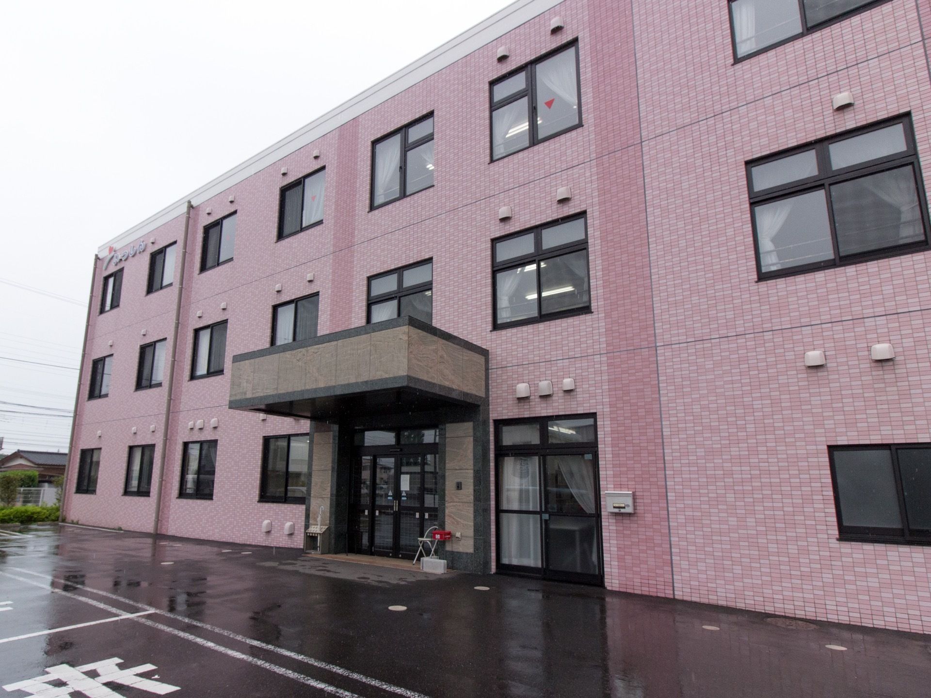 建物は壁がピンクのタイルが一面に貼られている。玄関前には屋根がせり出し、入口横にポストが設置されている。