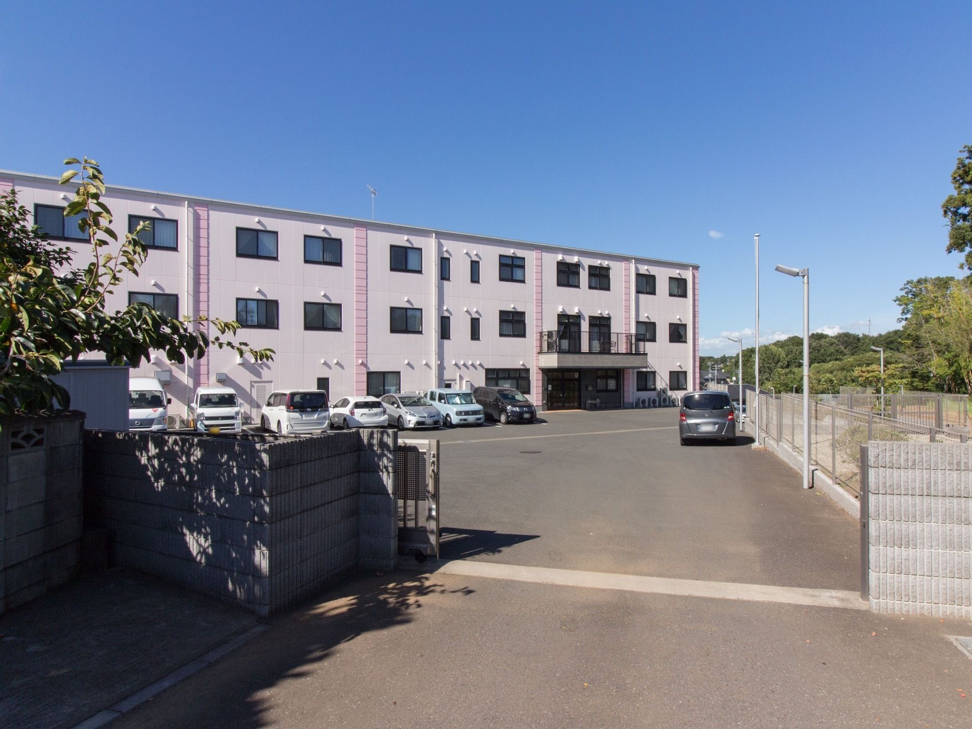 オフホワイトと薄いピンクに塗り分けられた外観で３階建てである。施設の前は駐車場になっている。鉄製の門がある。