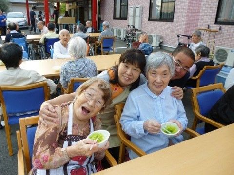 テーブルについて、手に小皿をもった高齢女性2人の肩を抱えるスタッフの女性