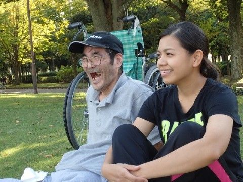 車椅子の前に腰を下ろす帽子をかぶった笑顔の高齢男性の右に膝を抱えて座る女性スタッフ