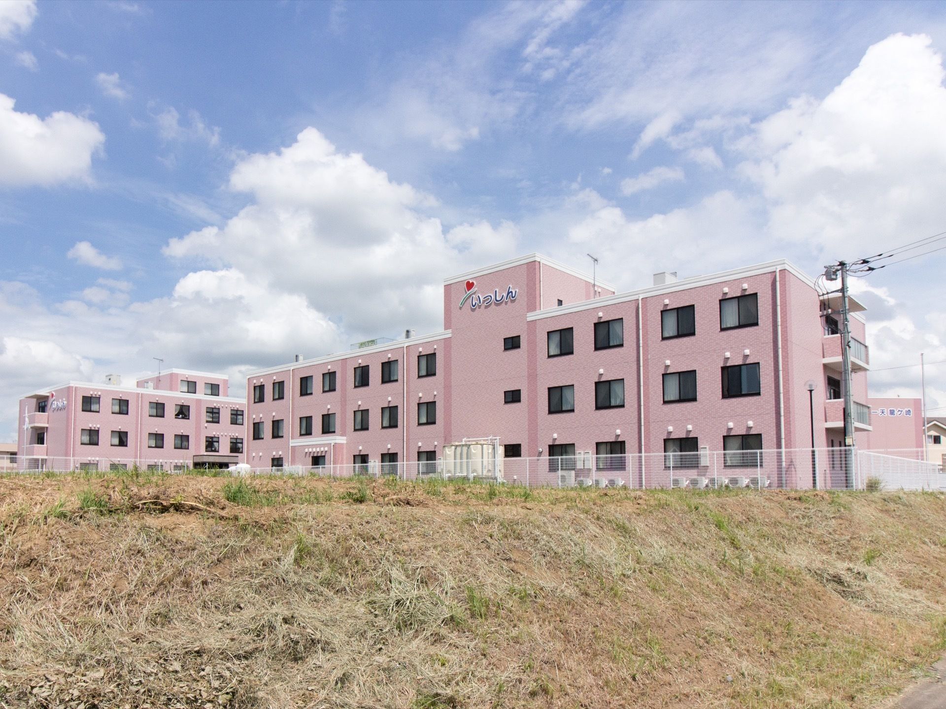 タイル貼りの明るいピンク色の外観が印象的な建物。壁面に施設名が大きく掲げられている。隣には似た造りで同じグループの施設が建っている。