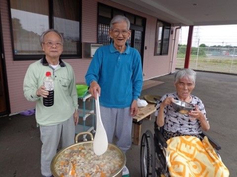 何かが煮えている大なべの後ろにしょうゆやしゃもじを持って立つ2人の高齢者とその横にいる車椅子を利用する高齢者