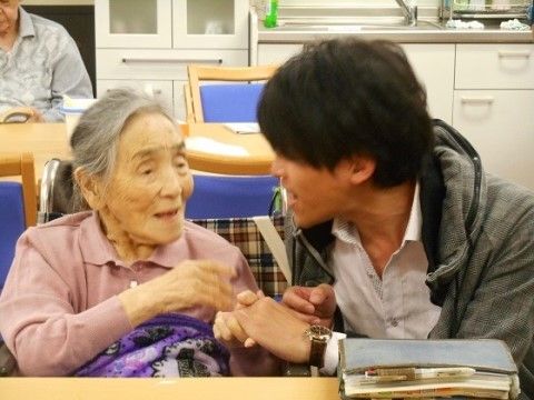 テーブルについたグレーがかったピンクのシャツを着た高齢女性の右手を取って話しかけるスタッフ