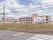 ピンク色の３階建の建物である。上には空が大きく広がっており、回りには高い建物が見当たらない。白いフェンスで敷地が囲まれている。