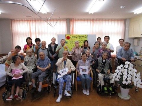 掲示物を貼ったホワイトボードの前で、車椅子や椅子に座った人を前列にして並ぶ入居者とスタッフ