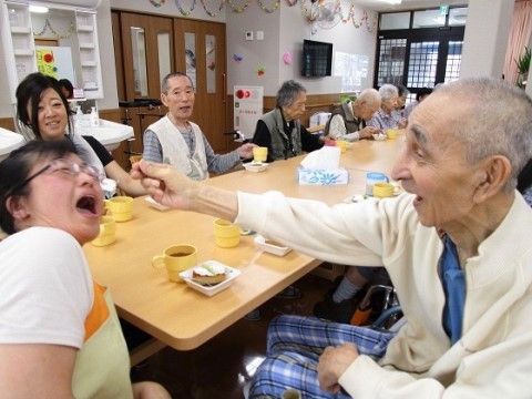 チェックのパジャマに白い上着の高齢男性が差し出したものを、口で捉えようとするスタッフ