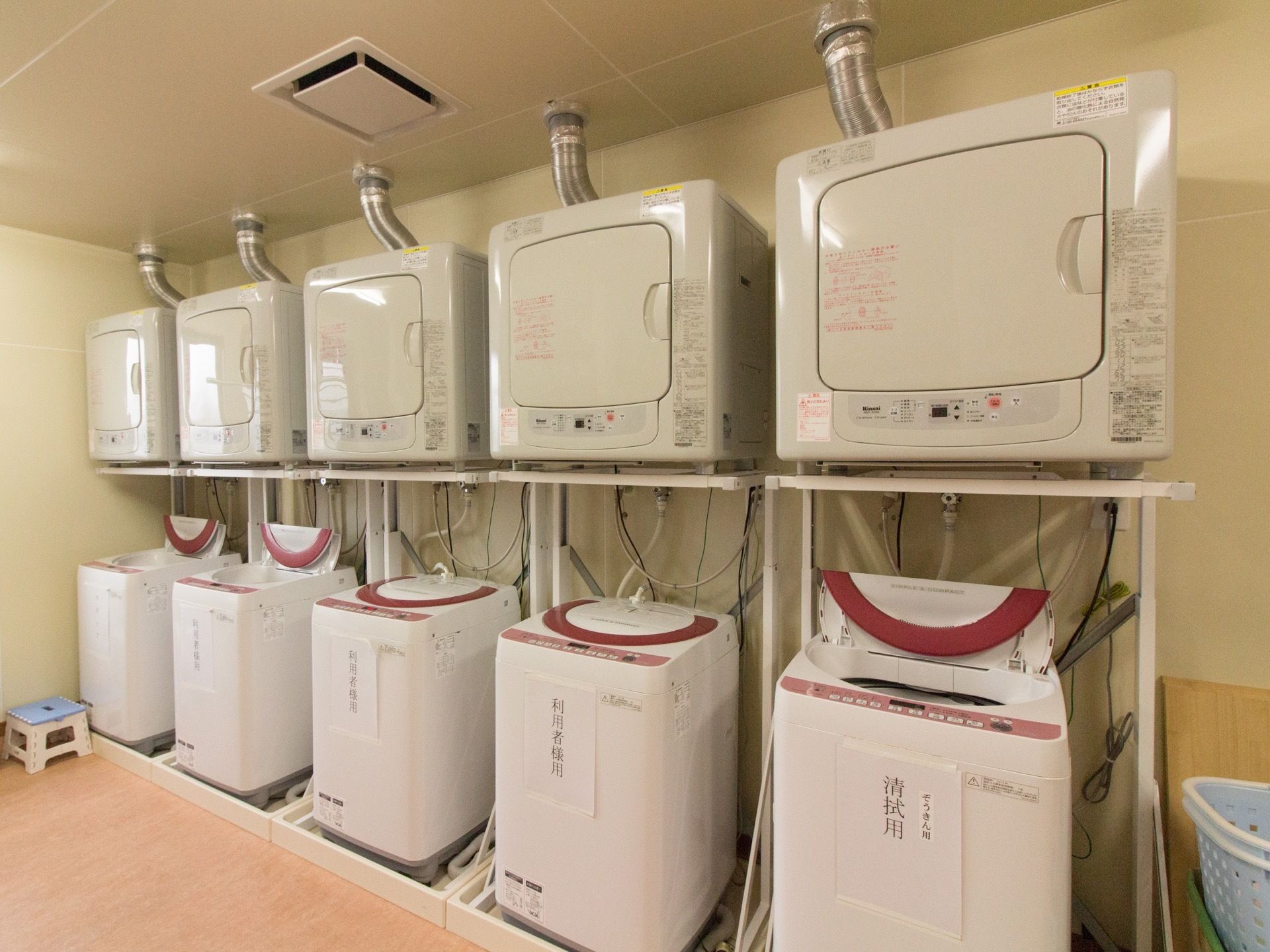 縦型の洗濯機、乾燥機が５台備えつけられたランドリールームである。水色のバスケットや踏み台も置かれている。